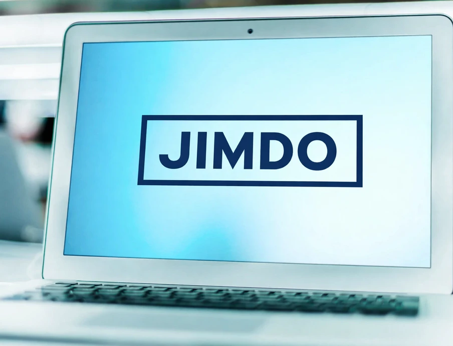 Laptop mit geöffnetem Bildschirm; Anzeige zeigt JIMDO in einem schwarzen Rechteck auf blauem Hintergrund.