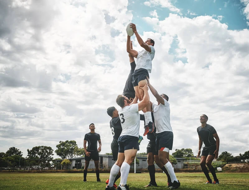 Rugbyspieler heben einen Mitspieler hoch, um den Ball zu fangen; weitere Spieler stehen auf dem Spielfeld.