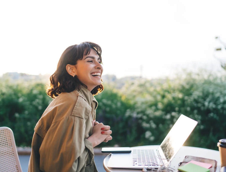 Eine Person sitzt im Freien vor einem Laptop; Smartphone, Notizbuch und Kaffeebecher auf dem Tisch, grüne Hecke dahinter.