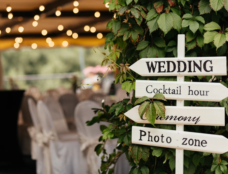 Holz-Wegweiser mit Pfeilen: WEDDING, Cocktail hour, Ceremony, Photo zone; Pflanze im Vordergrund, Tische im Hintergrund.