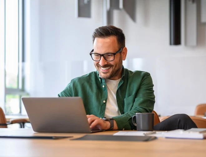 Mann mit Brille in grünem Hemd sitzt am Tisch vor Laptop, Kaffeebecher und Unterlagen; lächelt in hellem Büroraum.