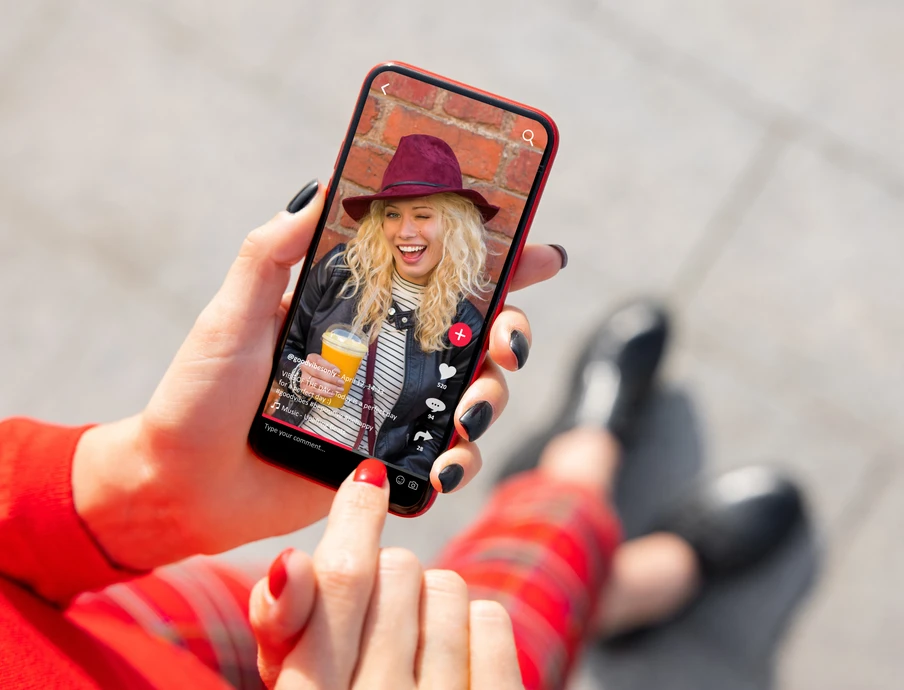 Auf dem Smartphonebildschirm eine blonde Frau mit violettem Hut, Schal und orangefarbenem Getränk vor Backsteinwand.
