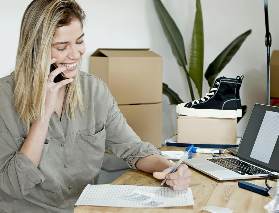 Eine Frau sitzt an einem Tisch, telefoniert und schreibt; Laptop, schwarzer Sneaker auf Box, Kartons und Pflanze im Hintergrund.