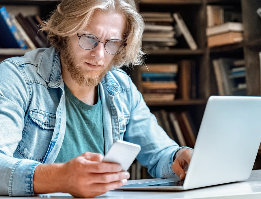 Mann mit Brille und Bart sitzt am Tisch, schaut auf Smartphone, Laptop davor; Bücherregale im Hintergrund.