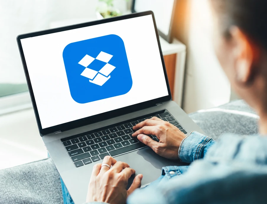 Person tippt auf Laptop; Bildschirm zeigt blaues Dropbox-Logo mit weißem Diamantsymbol.