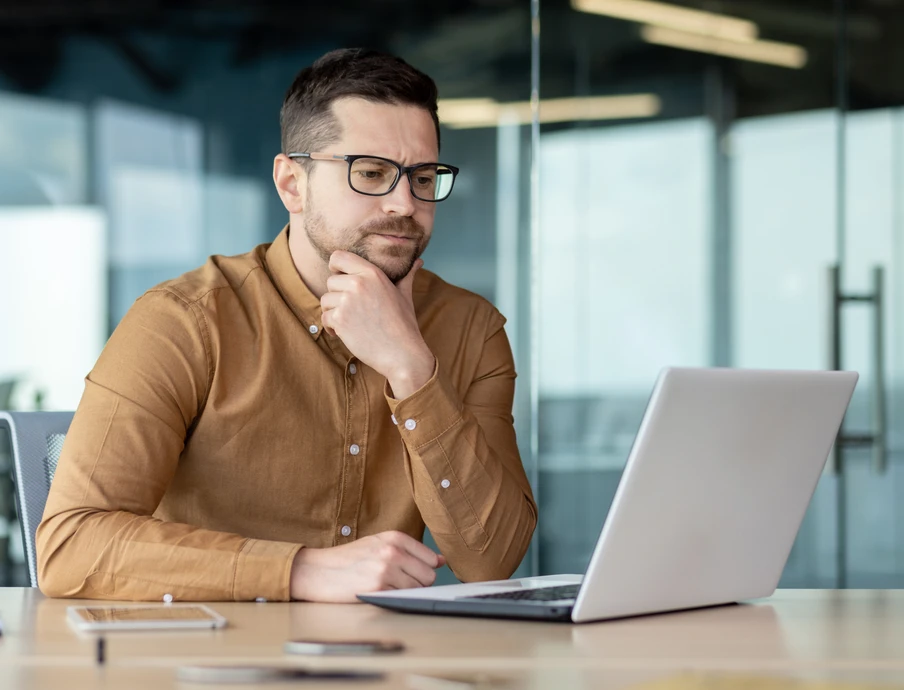 Mann mit Brille und braunem Hemd sitzt am Schreibtisch vor Laptop, Hand am Kinn, Büro mit Glaswänden.