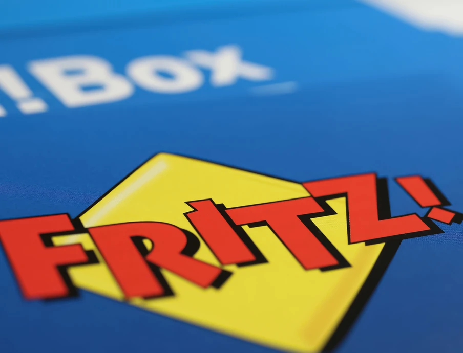 Nahaufnahme eines blauen Kartons mit gelbem Diamant-Logo und roter Schrift 'FRITZ!'; im Hintergrund teils weißer Text.