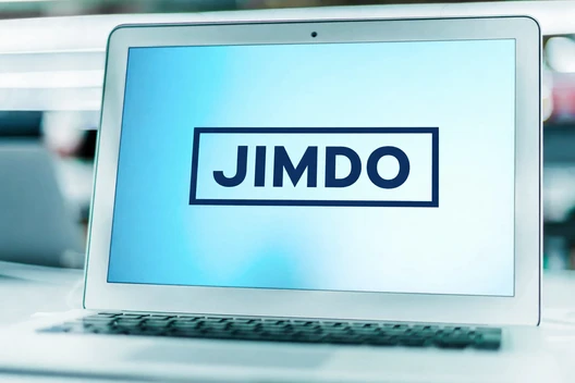 Laptop mit geöffnetem Bildschirm; Anzeige zeigt JIMDO in einem schwarzen Rechteck auf blauem Hintergrund.