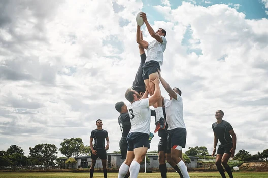 Rugbyspieler heben einen Mitspieler hoch, um den Ball zu fangen; weitere Spieler stehen auf dem Spielfeld.