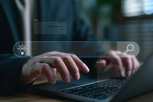 Hände tippen auf einem Laptop; eine blaue holografische Suchleiste mit Such-Icon erstreckt sich über die Tastatur.