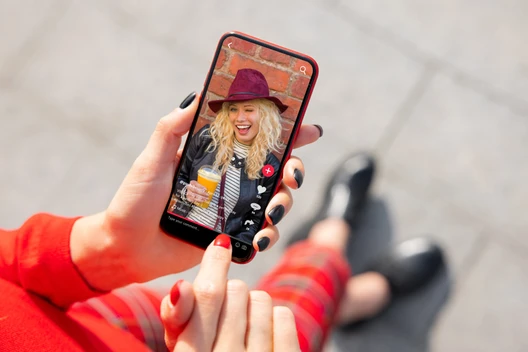 Auf dem Smartphonebildschirm eine blonde Frau mit violettem Hut, Schal und orangefarbenem Getränk vor Backsteinwand.