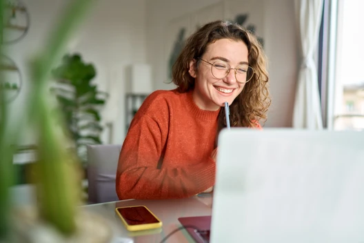Person mit Brille und orangefarbenem Pullover sitzt am Laptop, lächelt und hält Stift an den Mund.