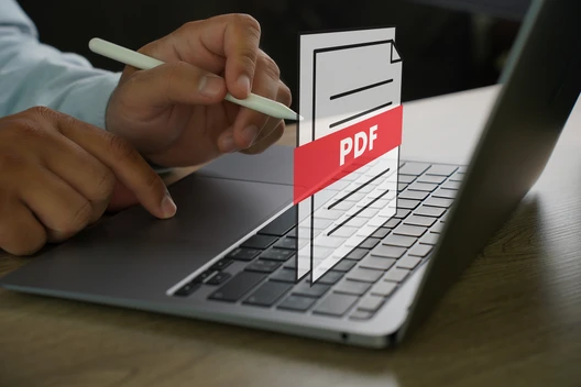 Nahaufnahme einer Hand mit Stylus über einer Laptop-Tastatur; davor ein 3D-Dokument-Icon mit rotem PDF-Band.