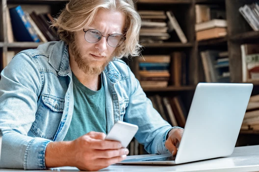 Mann mit Brille und Bart sitzt am Tisch, schaut auf Smartphone, Laptop davor; Bücherregale im Hintergrund.