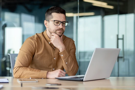 Mann mit Brille und braunem Hemd sitzt am Schreibtisch vor Laptop, Hand am Kinn, Büro mit Glaswänden.