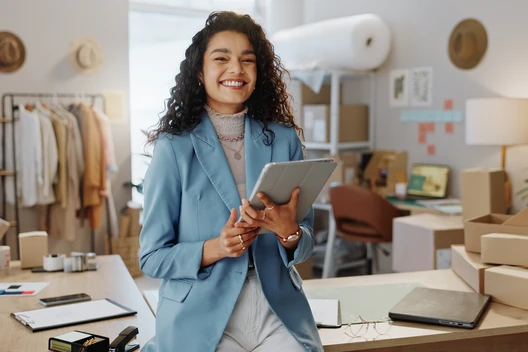 Eine Frau mit lockigen Haaren in blauem Blazer hält Tablet, lächelt; Hintergrund: Kartons, Laptop, Kleiderständer und Lampen.