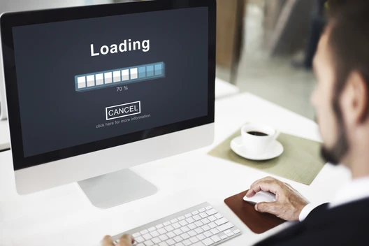iMac-Bildschirm zeigt Loading mit Fortschrittsbalken ca. 70%; weiße Tastatur, Maus, Kaffeetasse, Person rechts.