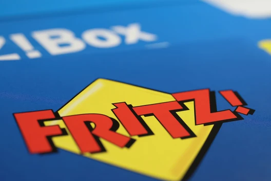 Nahaufnahme eines blauen Kartons mit gelbem Diamant-Logo und roter Schrift 'FRITZ!'; im Hintergrund teils weißer Text.